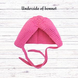 Puede incluir: Un gorro tejido a mano de color rosa brillante con un patr&oacute;n de punto bobo texturizado y dos lazos largos tejidos para atar. El texto "Underside of bonnet" est&aacute; impreso en negro sobre la prenda, mostrando la construcci&oacute;n interior del art&iacute;culo.