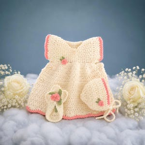 May include: A cream-coloured crochet baby dress set with pink trim, featuring a dress, bonnet, and booties. Each piece is adorned with small pink rose accents. The set is displayed on a soft, cloud-like surface.
