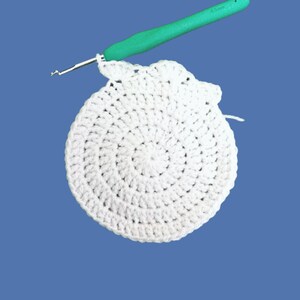Peut inclure: Cercle blanc crochet&eacute; en cours, avec un crochet vert en haut. Le crochet est ins&eacute;r&eacute; dans le fil blanc, montrant le d&eacute;but d'une nouvelle rang&eacute;e. Le fond est bleu uni. Le crochet est marqu&eacute; 4,5 mm.