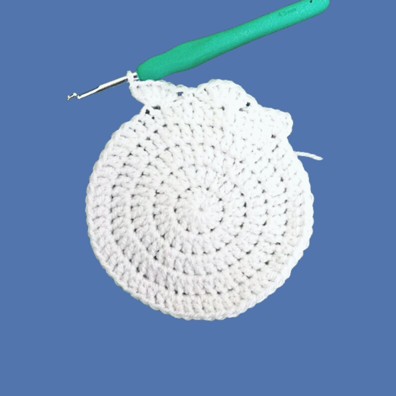 Peut inclure: Cercle blanc crochet&eacute; en cours, avec un crochet vert en haut. Le crochet est ins&eacute;r&eacute; dans le fil blanc, montrant le d&eacute;but d'une nouvelle rang&eacute;e. Le fond est bleu uni. Le crochet est marqu&eacute; 4,5 mm.