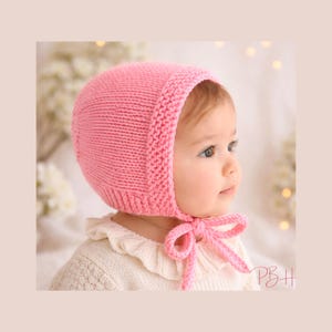 May include: A pink knitted baby bonnet with a rounded crown and a ribbed brim. The bonnet has a matching pink tie under the chin. The baby is wearing a white sweater with a ruffled collar.