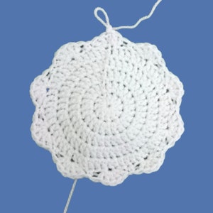 Peut inclure: Napperon blanc fait &agrave; la main au crochet, avec un bord festonn&eacute;. Le motif circulaire pr&eacute;sente un motif en spirale, avec une boucle en haut et un fil l&acirc;che. Le napperon est pos&eacute; sur un fond bleu.