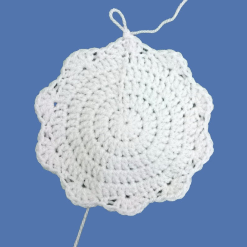 Peut inclure: Napperon blanc fait &agrave; la main au crochet, avec un bord festonn&eacute;. Le motif circulaire pr&eacute;sente un motif en spirale, avec une boucle en haut et un fil l&acirc;che. Le napperon est pos&eacute; sur un fond bleu.