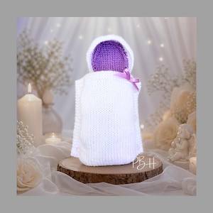 Könnte beinhalten: Handgestrickter weißer Baby-Bunting mit lila Kapuze und lavendelfarbener Schleife. Der Bunting wird auf einer Holzscheibe präsentiert, umgeben von Kerzen und Blumen, wodurch eine weiche, ruhige Atmosphäre entsteht.