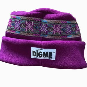 May include: A purple fleece beanie with a patterned band featuring geometric designs in teal, pink, and brown. The hat has a folded brim and a white rectangular tag with the text "DIGME JACKSON HOLE, WY".
