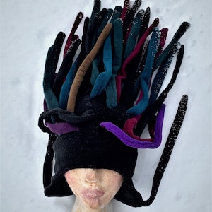 May include: A black fleece hat with multiple colorful, elongated fabric pieces extending upwards. The hat features purple, teal, brown, and burgundy accents, creating a unique and artistic design. The hat is displayed on a mannequin head.