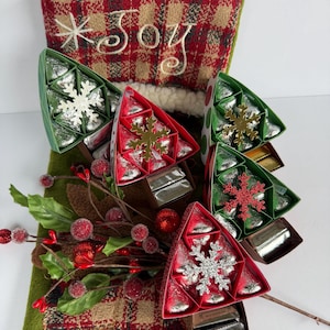 May include: A red and tan plaid Christmas stocking with the word Joy embroidered in white, featuring four triangular chocolate gift boxes shaped like trees, each topped with a snowflake ornament and decorated with red berry sprigs.