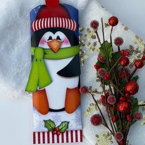 May include: A vertical rectangular decorative sign featuring a cartoon penguin wearing a red and white striped hat with a red pom-pom and a bright green scarf. The penguin has orange feet and beak, with a holly sprig and red striped border at the base.