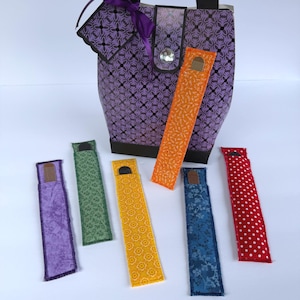 May include: A purple patterned tote bag with black handles, a central snap closure featuring a clear gem, and a purple ribbon accent. Six long, rectangular fabric bookmarks in various colors and patterns are arranged in front of the bag.