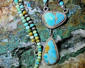 May include: A turquoise and silver necklace with a teardrop pendant. The necklace features turquoise beads in shades of blue, green, and yellow. The pendant has a silver rope-like border. The necklace is displayed on a textured, multi-colored stone surface.