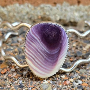 May include: A silver bracelet with a wavy design features a large, oval-shaped purple and white shell inlay.