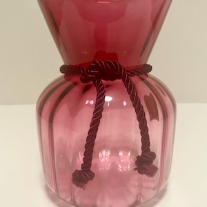 May include: A cranberry-colored glass vase with a bulbous base and a flared top. A dark red rope is tied around the middle of the vase. The vase has vertical ridges and a smooth, glossy finish.