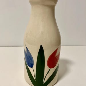 May include: Cream-colored ceramic vase with a milk bottle shape. The vase is decorated with a green stem and leaves, and red and blue tulip flowers. The vase has a smooth, glossy finish.