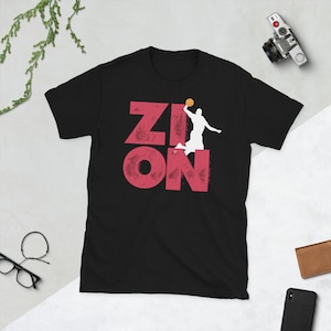 Pode incluir: Camiseta preta com a palavra "ZION" em letras vermelhas desgastadas e uma silhueta branca de um jogador de basquete. A bola de basquete é amarela. Uma camiseta casual para os fãs de esportes.