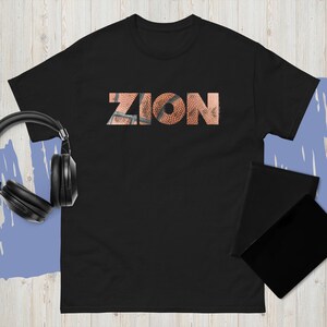 Pode incluir: Camiseta preta com a palavra "ZION" impressa no peito com textura de basquete. Fones de ouvido pretos e um tablet preto estão ao lado.