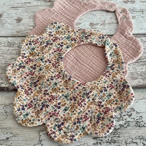 Könnte beinhalten: Zwei Baby-Lätzchen. Eines ist hellrosa, das andere ist weiß mit einem Blumenmuster aus rosa, blauen und gelben Blüten. Beide Lätzchen haben einen gewellten Rand und Druckknöpfe.
