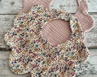Könnte beinhalten: Zwei Baby-Lätzchen. Eines ist hellrosa, das andere ist weiß mit einem Blumenmuster aus rosa, blauen und gelben Blüten. Beide Lätzchen haben einen gewellten Rand und Druckknöpfe.