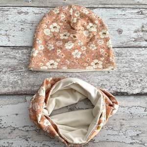 Może przedstawiać: Zestaw dwóch pasujących do siebie akcesoriów z tkaniny w kwiatowy wzór. Czapka typu beanie i komin są wykonane z tkaniny w kolorze brzoskwiniowym z białym nadrukiem kwiatowym.