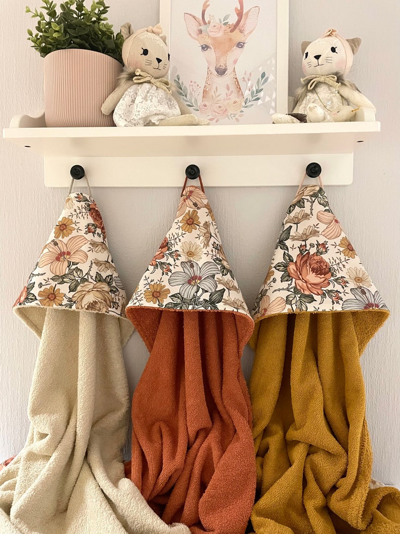 May include: Three hooded towels with floral patterns in shades of orange, yellow, and green. The towels are hanging on hooks and are made of a soft, absorbent material.