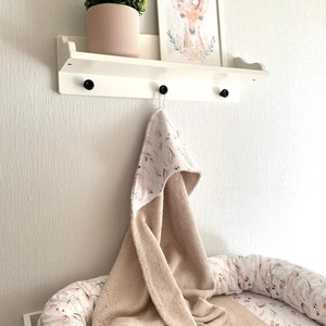 May include: A white shelf with a brown and white hooded towel hanging from a hook. The shelf has a small potted plant and a framed print of a deer with a floral wreath around its neck. The towel is on top of a white and brown patterned baby nest.
