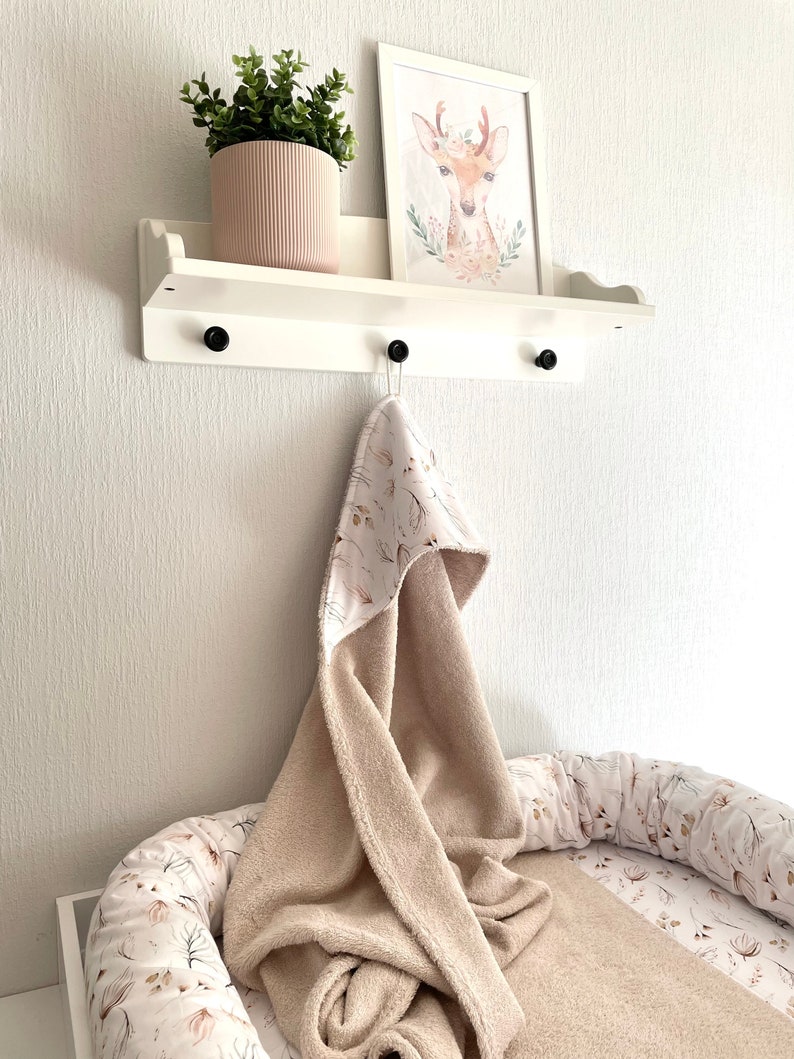 May include: A white shelf with a brown and white hooded towel hanging from a hook. The shelf has a small potted plant and a framed print of a deer with a floral wreath around its neck. The towel is on top of a white and brown patterned baby nest.