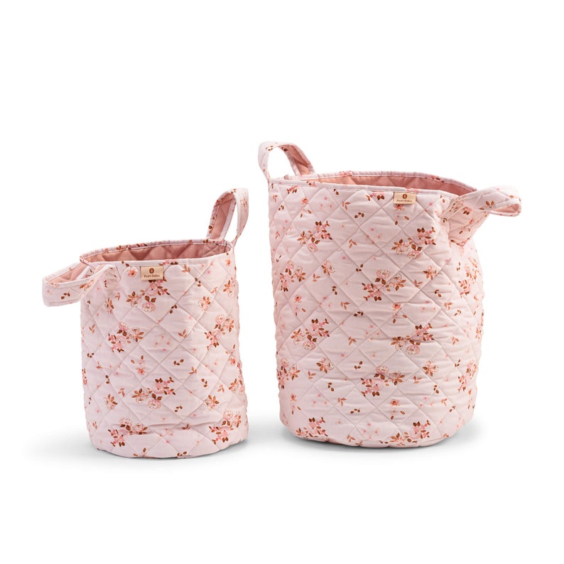 K&ouml;nnte beinhalten: Zwei rosa gesteppte Aufbewahrungsk&ouml;rbe mit Blumenmustern. Der gr&ouml;&szlig;ere Korb hat eine breite &Ouml;ffnung und einen einzelnen Griff. Der kleinere Korb hat eine schmalere &Ouml;ffnung und zwei Griffe.