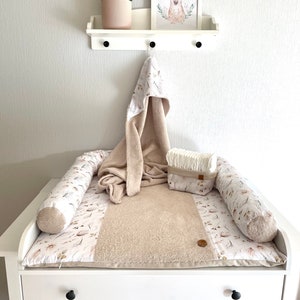 May include: A white changing table with two drawers and a beige changing pad with a floral pattern. The changing pad has two beige and white rolled cushions on either side. A white hooded towel and a white diaper pail are on top of the changing pad.