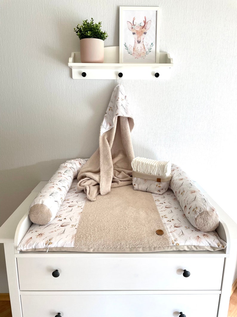 May include: A white changing table with two drawers and a beige changing pad with a floral pattern. The changing pad has two beige and white rolled cushions on either side. A white hooded towel and a white diaper pail are on top of the changing pad.