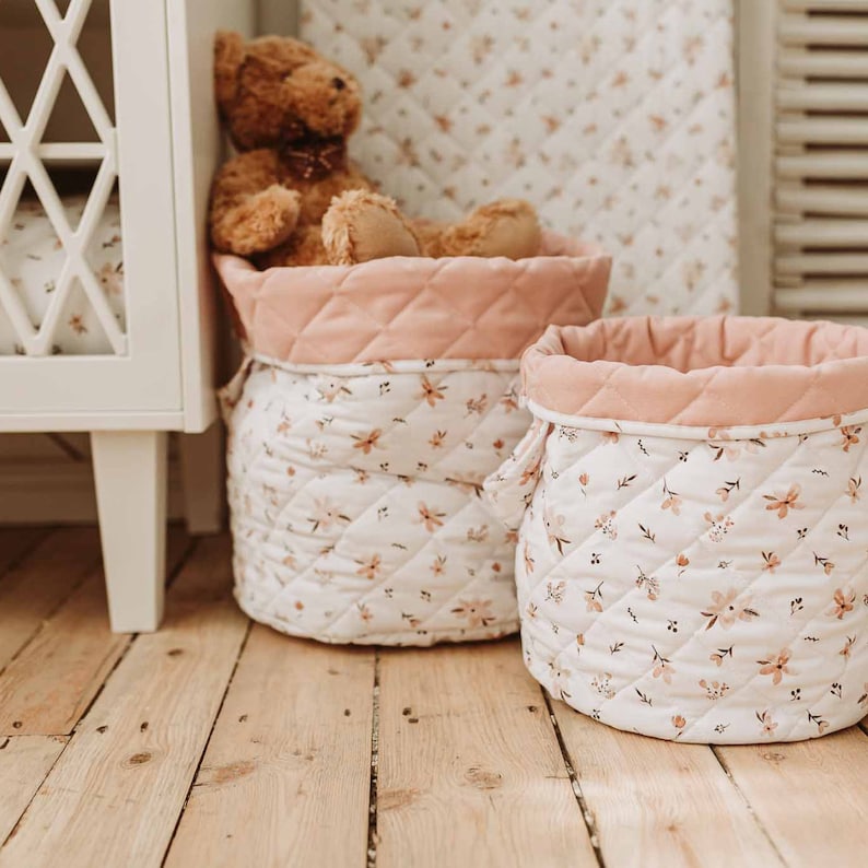 K&ouml;nnte beinhalten: Zwei gepolsterte Aufbewahrungsk&ouml;rbe mit rosa Besatz und einem Blumenmuster. Die K&ouml;rbe stehen auf einem Holzboden.