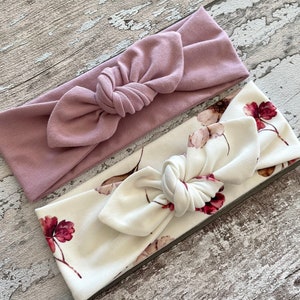 May include: Two fabric headbands, one is a solid dusty rose color and the other is white with a floral pattern in shades of pink and burgundy.