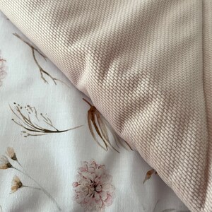 May include: A close-up of a soft, pale pink fabric with a textured surface. The fabric is folded over a white background with a delicate floral pattern featuring brown branches and pink flowers.