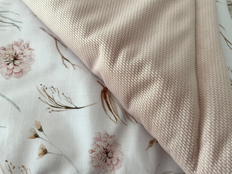 May include: A close-up of a soft, pale pink fabric with a textured surface. The fabric is folded over a white background with a delicate floral pattern featuring brown branches and pink flowers.