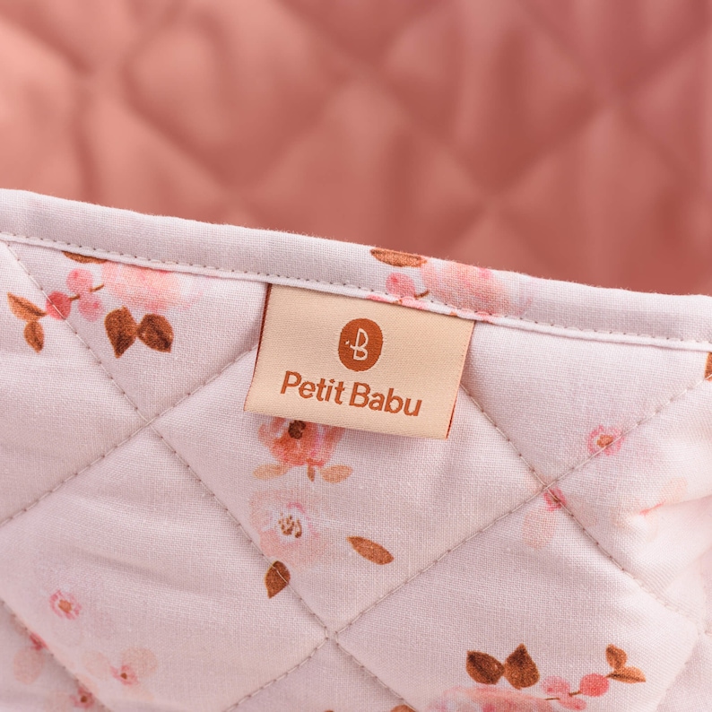 K&ouml;nnte beinhalten: Ein rosa-wei&szlig;er gesteppter Aufbewahrungskorb mit einem braunen Etikett, auf dem "Petit Babu" mit einem kleinen "B"-Logo steht.