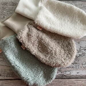 May include: Three pairs of soft, plush baby mittens in white, beige, and light green. The mittens are made of a soft, absorbent material and are perfect for keeping baby's hands warm and dry.