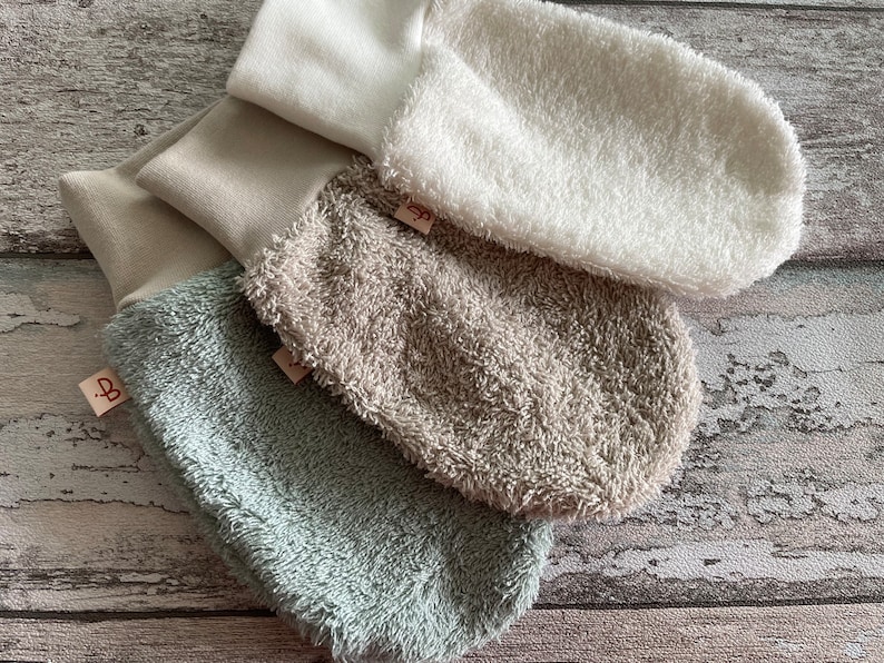 May include: Three pairs of soft, plush baby mittens in white, beige, and light green. The mittens are made of a soft, absorbent material and are perfect for keeping baby's hands warm and dry.
