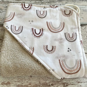Peut inclure: Une serviette &agrave; capuche avec un dos en &eacute;ponge beige et un devant en coton blanc orn&eacute; d'un motif r&eacute;p&eacute;t&eacute; d'arcs-en-ciel bruns, roses et gris.