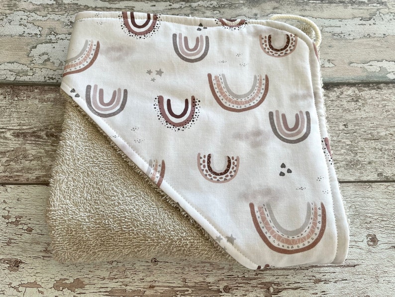 Peut inclure: Une serviette &agrave; capuche avec un dos en &eacute;ponge beige et un devant en coton blanc orn&eacute; d'un motif r&eacute;p&eacute;t&eacute; d'arcs-en-ciel bruns, roses et gris.