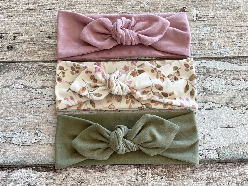 May include: Three knotted headbands in different colours and patterns. The first headband is a solid dusty rose colour. The second headband is a white fabric with a brown and pink floral pattern. The third headband is a solid olive green colour.