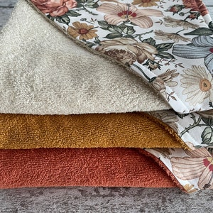 May include: A stack of four hooded towels with a floral print. The top towel is white with a beige floral pattern, the second towel is mustard yellow, the third towel is terracotta red, and the bottom towel is white with a beige floral pattern.
