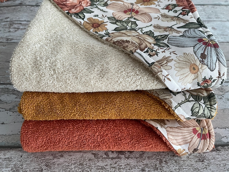 May include: A stack of four hooded towels with a floral print. The top towel is white with a beige floral pattern, the second towel is mustard yellow, the third towel is terracotta red, and the bottom towel is white with a beige floral pattern.