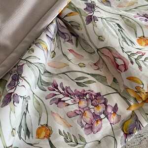 May include: White fabric with a floral print featuring purple, yellow, and pink flowers. The fabric has a subtle sheen and a soft texture.