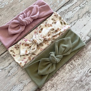 May include: A set of three fabric headbands. One is a solid dusty rose colour, one is a white headband with a brown floral pattern, and the last is a solid olive green headband. All headbands are tied in a knot at the top.