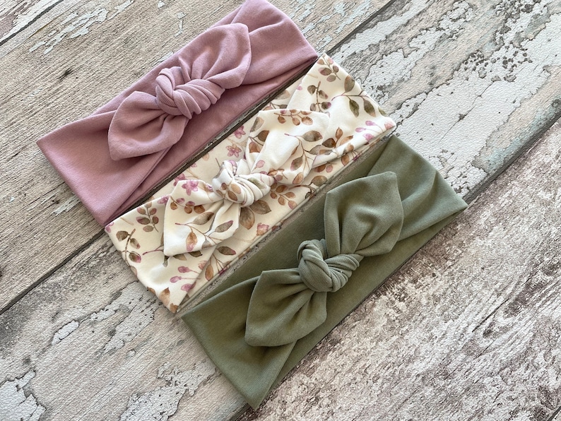 May include: A set of three fabric headbands. One is a solid dusty rose colour, one is a white headband with a brown floral pattern, and the last is a solid olive green headband. All headbands are tied in a knot at the top.