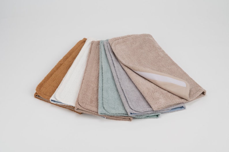 K&ouml;nnte beinhalten: Ein Set aus sieben saugf&auml;higen Wickelunterlagen in verschiedenen Farben, darunter Braun, Wei&szlig;, Beige, Blau und Grau. Die Unterlagen bestehen aus weichem, pl&uuml;schem Stoff und haben eine wasserdichte R&uuml;ckseite.