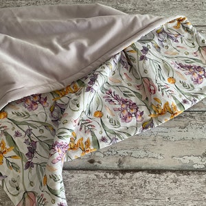 May include: A soft, light purple blanket with a floral print on the underside. The floral print features a white background with yellow, purple, and pink flowers.