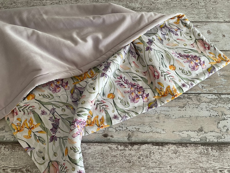 May include: A soft, light purple blanket with a floral print on the underside. The floral print features a white background with yellow, purple, and pink flowers.