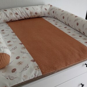 May include: A white changing pad with a brown waffle-textured insert and a white bolster with a floral print and brown accents. The bolster is placed around the perimeter of the changing pad.