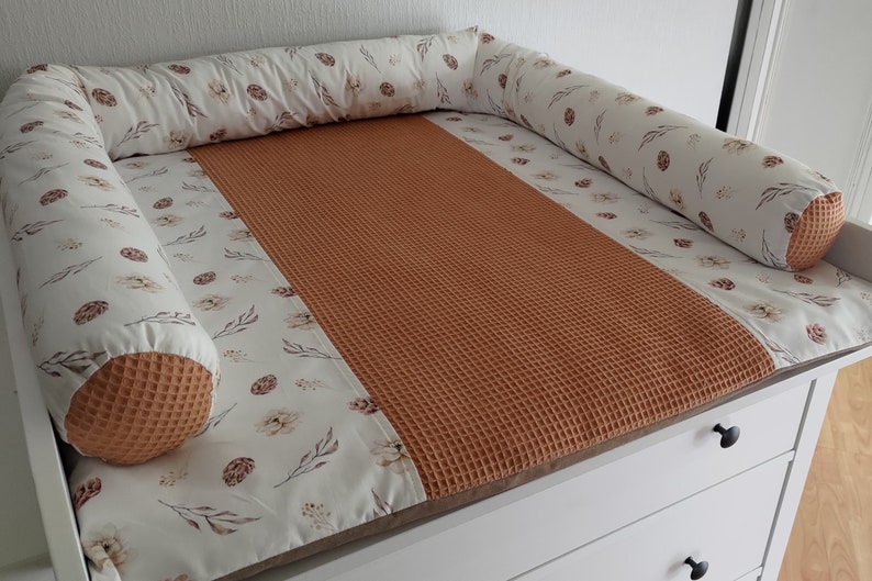 May include: A white changing pad with a brown waffle-textured insert and a white bolster with a floral print and brown accents. The bolster is placed around the perimeter of the changing pad.