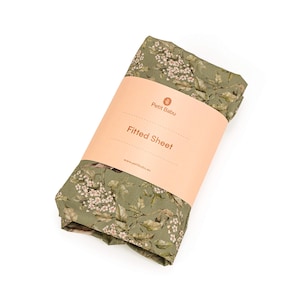 Puede incluir: Una sábana bajera verde con estampado floral con flores blancas y pájaros pequeños. La sábana está enrollada y tiene una etiqueta que dice "Petit Babu Sábana bajera".