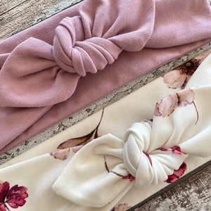 May include: Two fabric headbands, one is a solid dusty rose color and the other is white with a floral print. Both headbands are tied in a knot at the top.